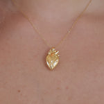 A gold anatomical heart pendant necklace worn on a person's neck, shown against light skin with a black bar at the bottom of the frame.