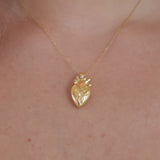 A gold anatomical heart pendant necklace worn on a person's neck, shown against light skin with a black bar at the bottom of the frame.
