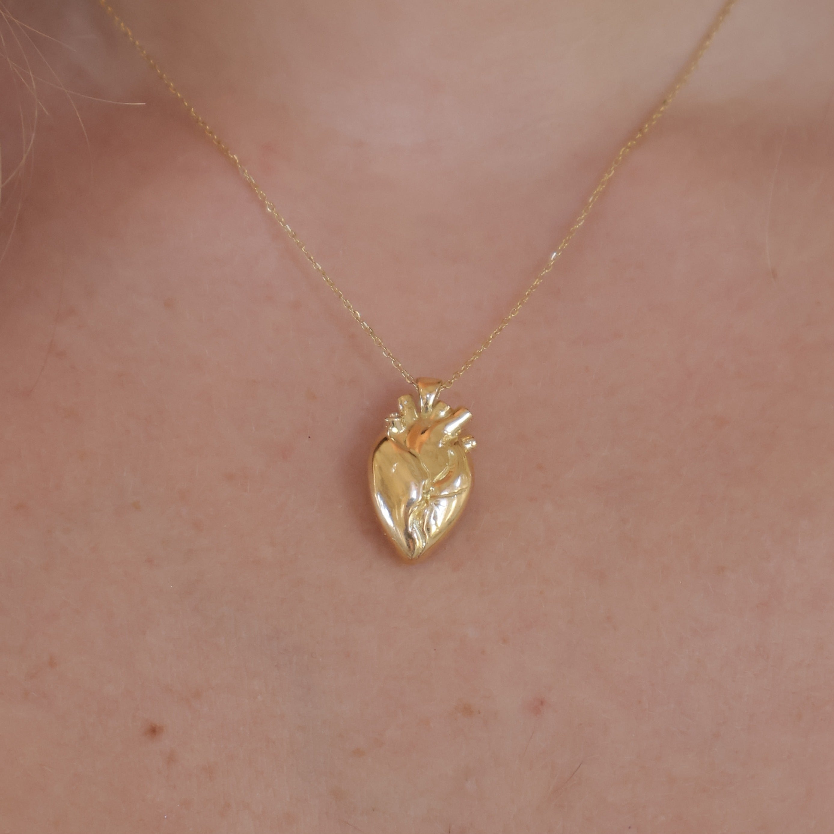 A gold anatomical heart pendant necklace worn on a person's neck, shown against light skin with a black bar at the bottom of the frame.