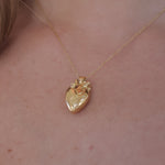 A close-up of a delicate gold necklace with a textured heart-shaped pendant, worn on a person's neck.