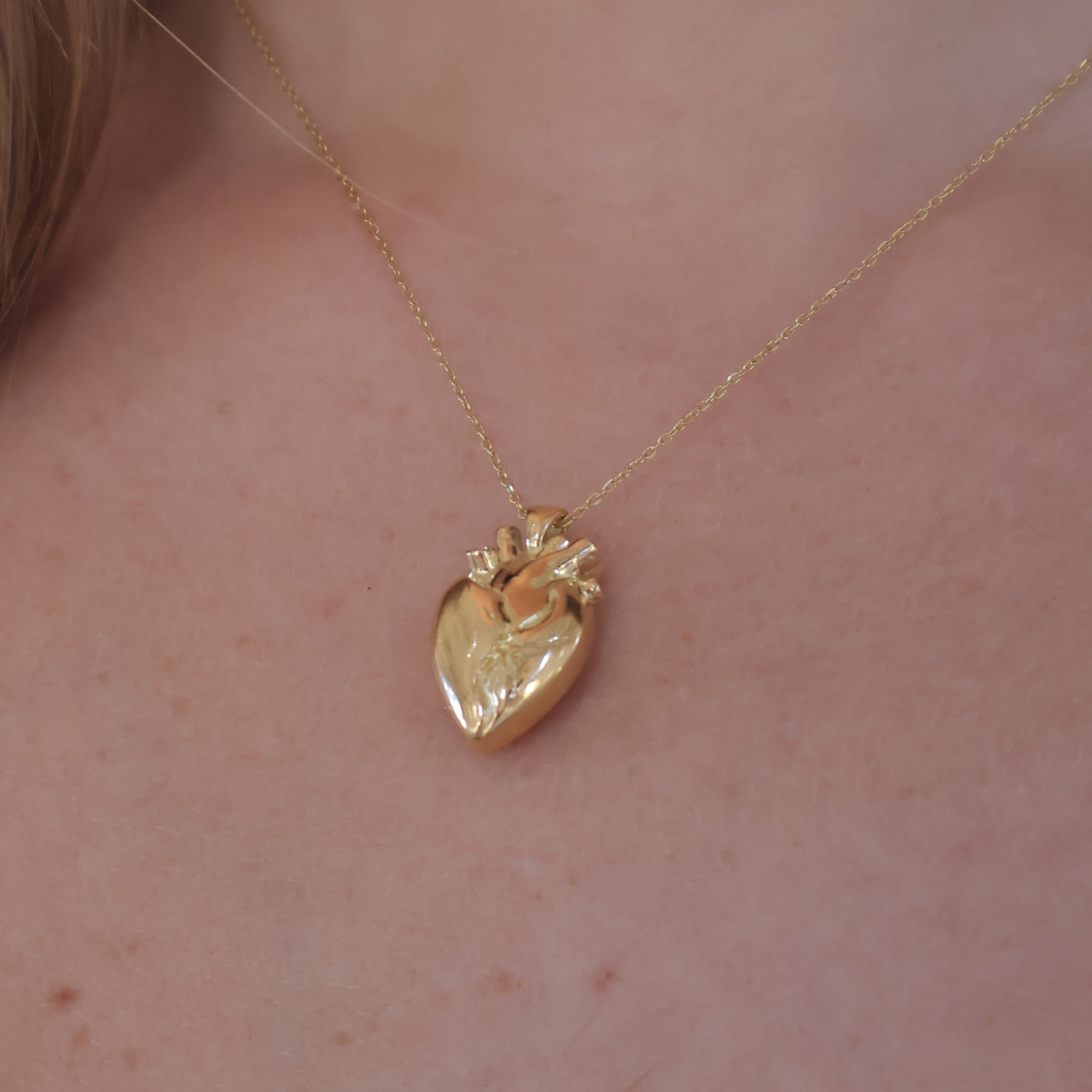 A close-up of a delicate gold necklace with a textured heart-shaped pendant, worn on a person's neck.