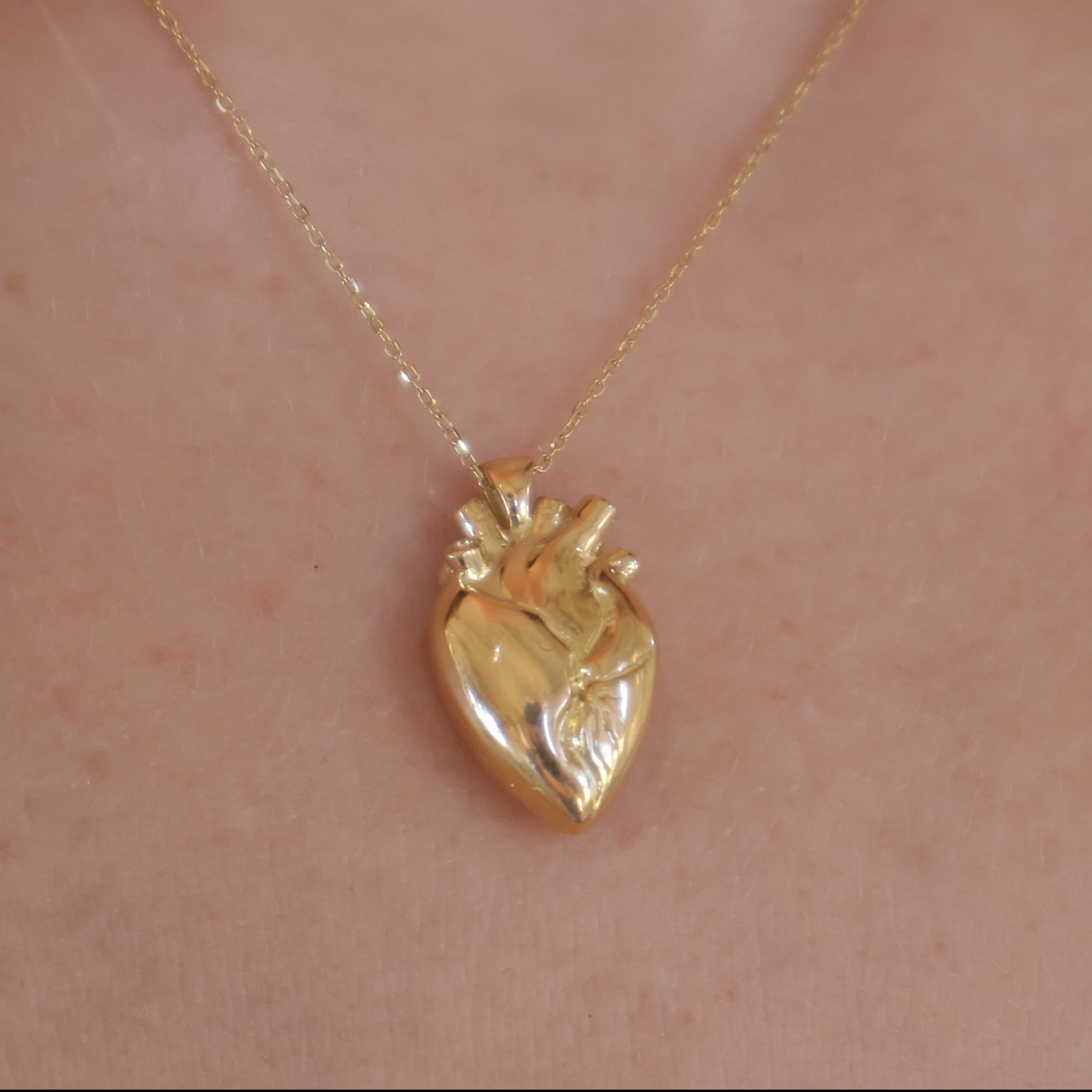 A gold anatomical heart pendant necklace worn on a person's neck, shown against light skin.