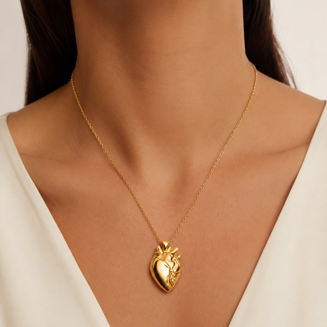 A gold anatomical heart pendant necklace worn by a model in a close-up shot. The model is wearing a white V-neck top, and the background is a soft, neutral beige.
