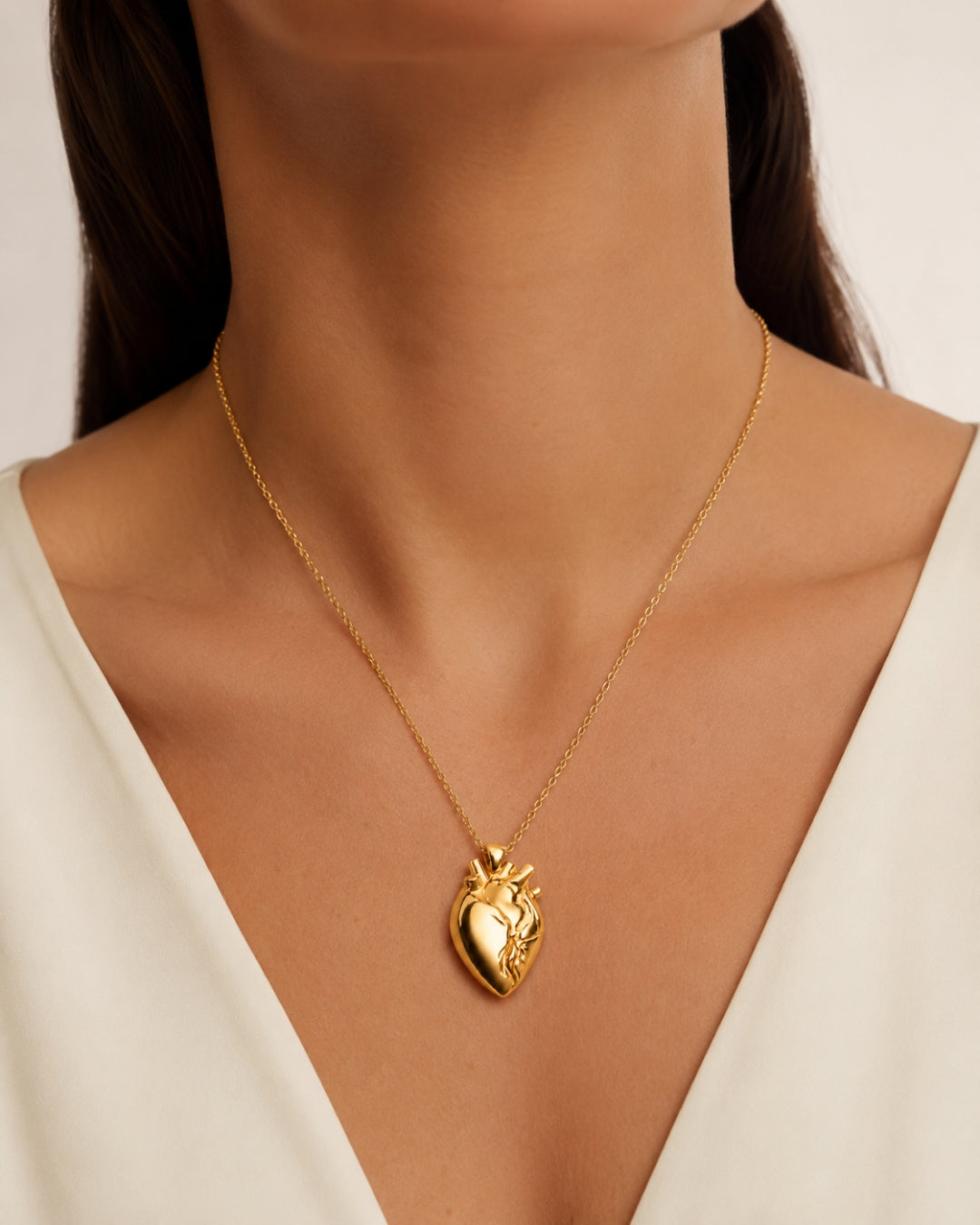 A gold anatomical heart pendant necklace worn by a model in a close-up shot. The model is wearing a white V-neck top, and the background is a soft, neutral beige.