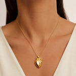 A gold anatomical heart pendant necklace worn by a model in a close-up shot. The model is wearing a white V-neck top, and the background is a soft, neutral beige.