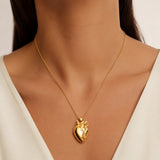 A gold anatomical heart pendant necklace worn by a model in a close-up shot. The model is wearing a white V-neck top, and the background is a soft, neutral beige.
